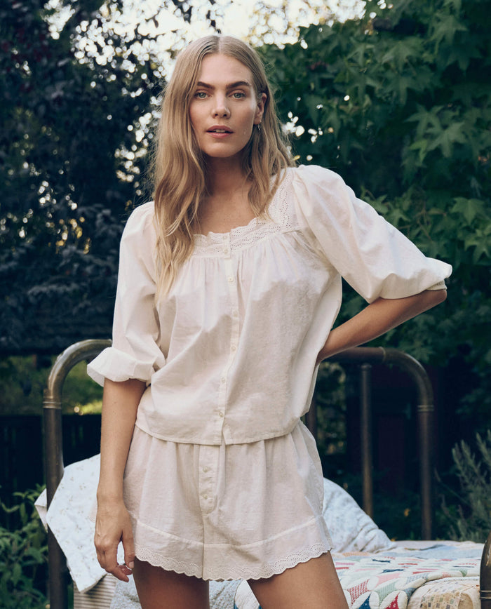 A woman stands outdoors against greenery, wearing THE GREAT. CORE SLEEP's The Eyelet Tap Short in True White with a loose, light-colored cotton voile blouse.