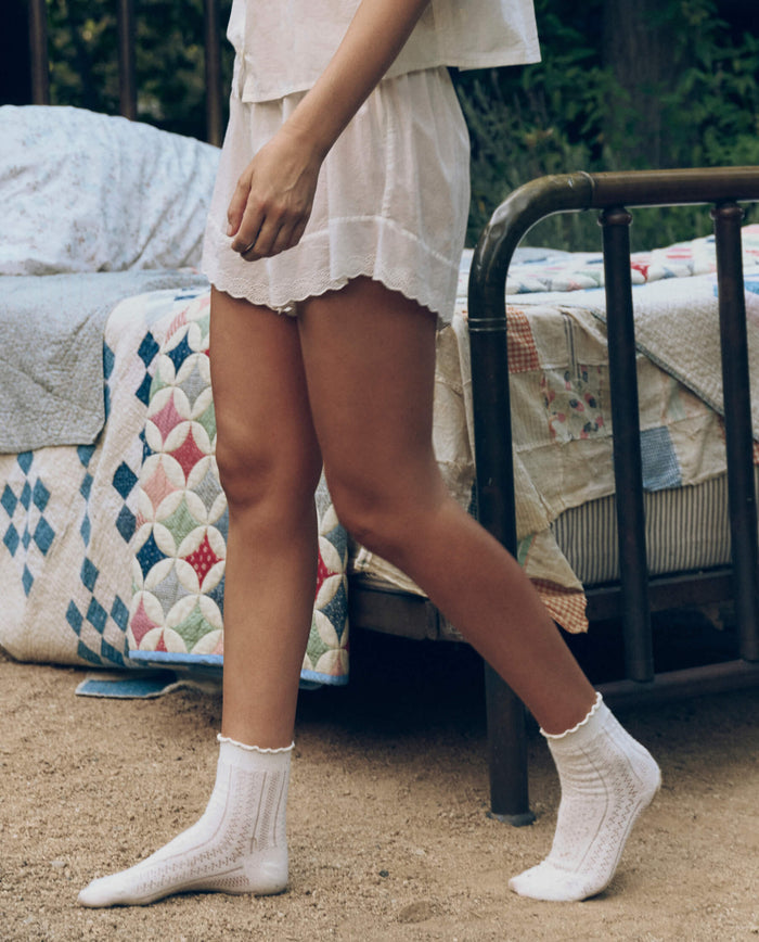 Wearing THE GREAT. CORE SLEEP’s Eyelet Tap Short in True White, a person with socks stands beside an outdoor bed with a metal frame and colorful quilt, set on a dirt surface.