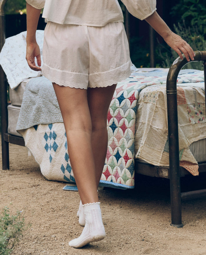 Someone in THE GREAT. CORE SLEEP's Eyelet Tap Short in True White and socks walks toward a metal bed with colorful patchwork quilts, outdoors on dirt ground.