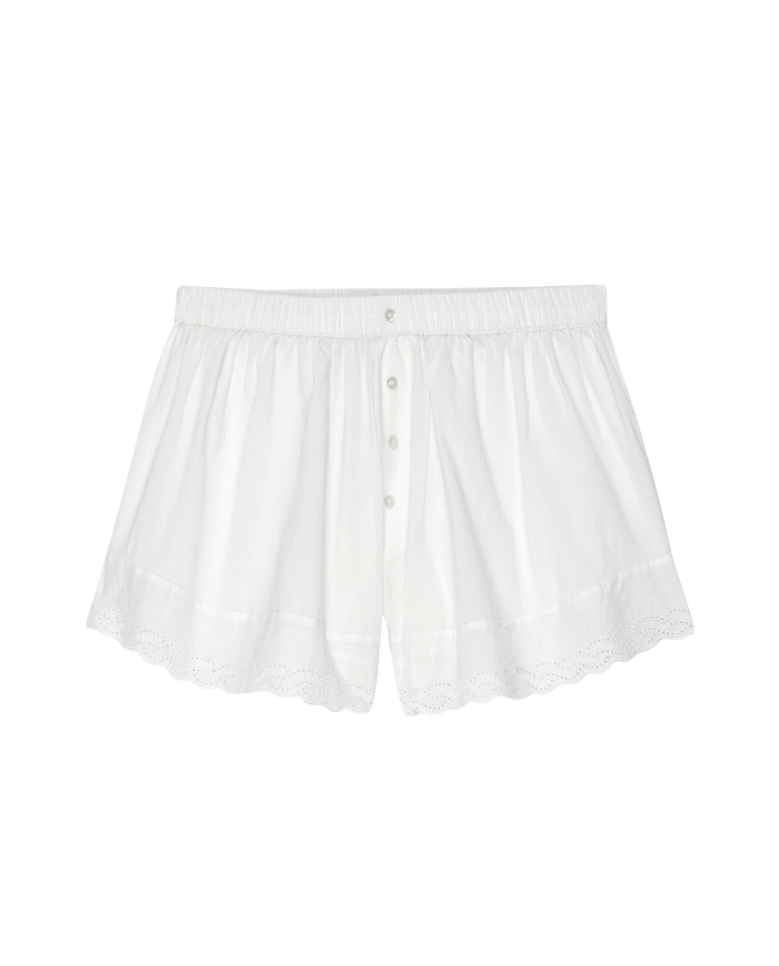 The Eyelet Tap Short in True White by THE GREAT. PS25 SLEEP, features a lightweight cotton voile material with scalloped hem, eyelet lace trim, elastic waistband, and button front closure, displayed on a black background.