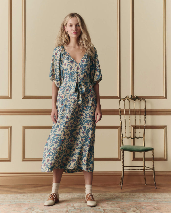 A woman stands indoors in THE GREAT. SP26 D2’s The Fairground Dress – Blue Scattered Bloom, featuring a belt and short sleeves. She pairs it with ankle socks and tan lace-up shoes; a green cushioned chair sits beside her.