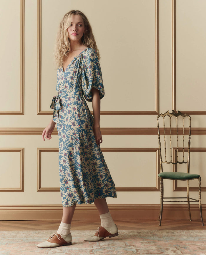A woman in THE GREAT. SP26 D2's The Fairground Dress – Blue Scattered Bloom walks past an ornate chair in a paneled room, wearing brown shoes with socks.