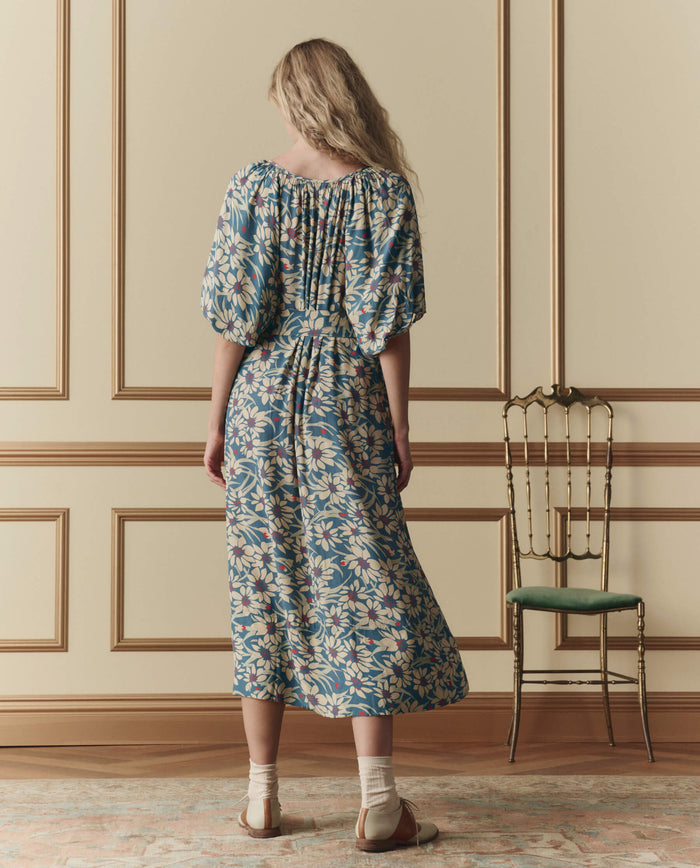 A woman stands indoors facing away, wearing THE GREAT. SP26 D2 'The Fairground Dress' in Blue Scattered Bloom. She pairs it with sandals and socks beside a green-seated chair against a beige paneled wall.