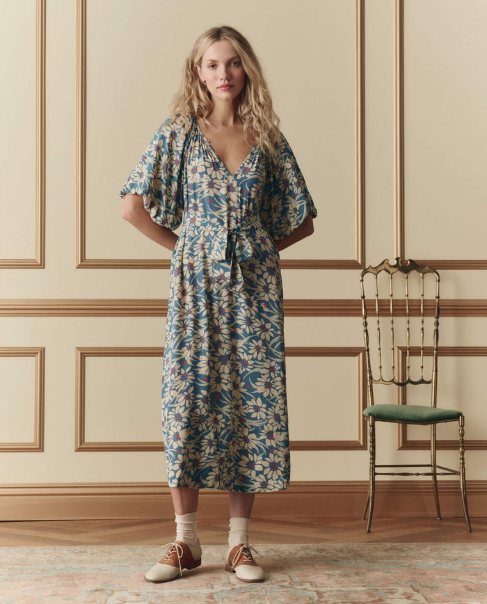A woman stands indoors in THE GREAT. SP26 D2 The Fairground Dress – Blue Scattered Bloom, paired with white socks and brown-and-white shoes, beside a green cushioned chair against a paneled wall.