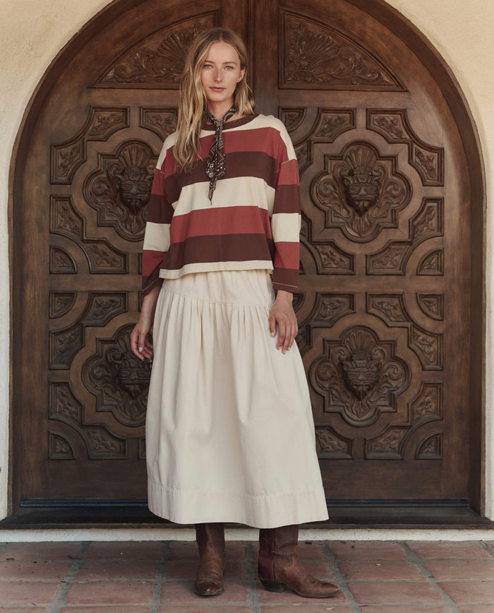 A woman stands in front of an ornate wooden door, wearing THE GREAT. PF26 The Field Tee -- Ashwood Stripe beneath a scarf, paired with a cream-colored skirt and brown boots.