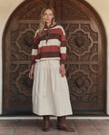 A woman stands in front of an ornate wooden door, wearing THE GREAT. PF26 The Field Tee -- Ashwood Stripe beneath a scarf, paired with a cream-colored skirt and brown boots.
