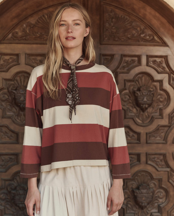 A woman stands before an ornate wooden door, wearing THE GREAT. PF26 The Field Tee in Ashwood Stripe, paired with a cream skirt and a patterned neck scarf.