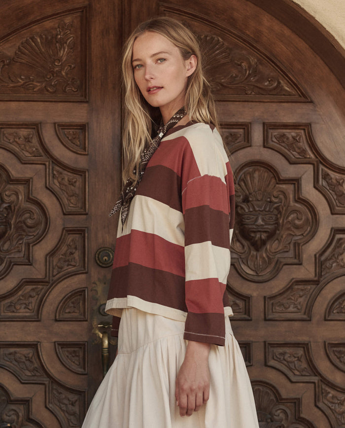 A woman stands before an ornate wooden door, wearing THE GREAT. PF26's The Field Tee in Ashwood Stripe, paired with a cream skirt and a neck scarf, looking toward the camera.
