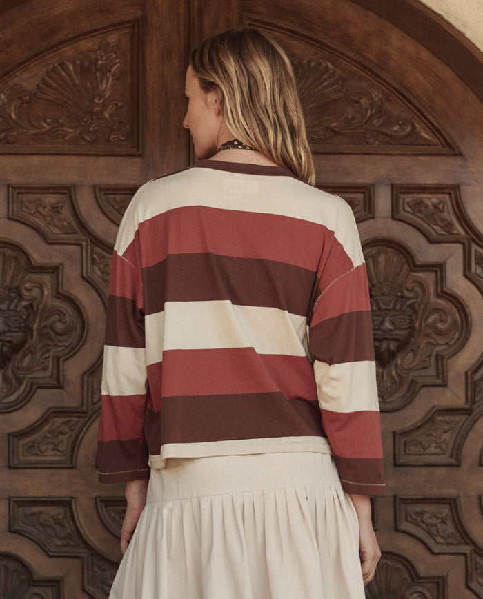 Seen from behind, a person with long hair stands before an ornate wooden door, wearing THE GREAT. PF26 The Field Tee in Ashwood Stripe—a loose brown and cream striped top—paired with a light-colored skirt.