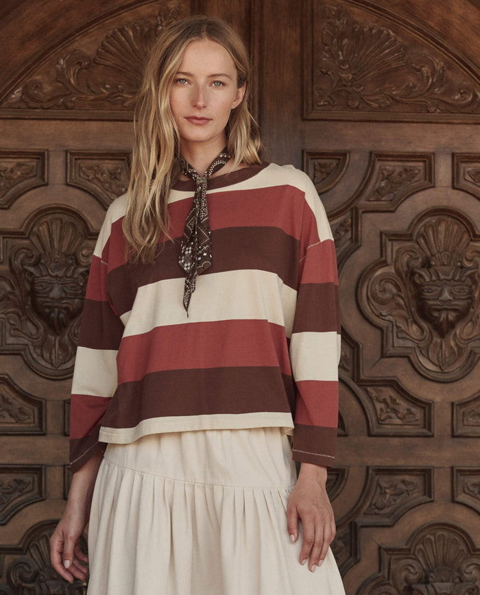 A woman stands before an ornate wooden door, wearing THE GREAT. PF26 The Field Tee in Ashwood Stripe, a cream skirt, and a patterned neck scarf.
