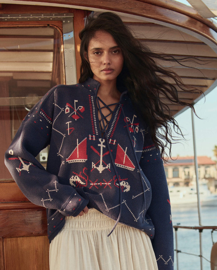 A person stands on a boat in THE GREAT. SU26's The Fisherman Laceup Pullover in navy, paired with a white skirt; water and buildings are visible in the background.