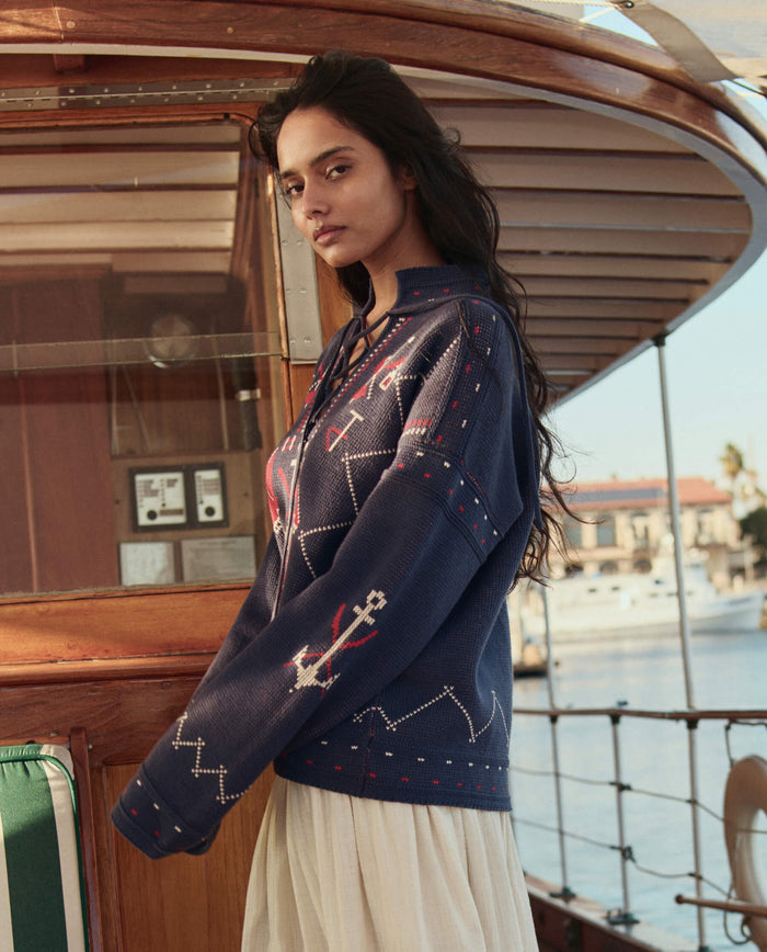 A woman stands on a boat in THE GREAT. SU26 The Fisherman Laceup Pullover in navy, featuring white patterns, paired with a cream skirt. Water and buildings are visible in the background.