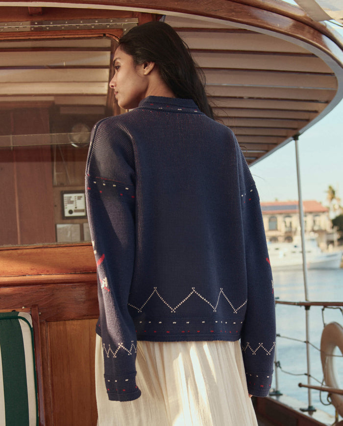 A woman with long dark hair, wearing THE GREAT. SU26 The Fisherman Laceup Pullover in navy, stands on a wooden boat facing away from the camera.