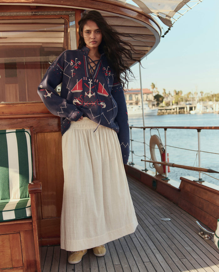A woman stands on a wooden boat deck wearing THE GREAT. SU26 The Fisherman Laceup Pullover in navy and a long beige skirt, with water and buildings in the background.