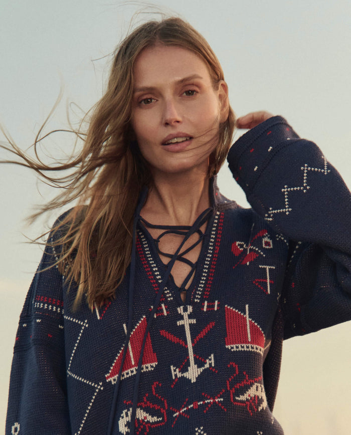 A woman with long hair stands outdoors in the wind, wearing THE GREAT. SU26 The Fisherman Laceup Pullover in navy, featuring red and white nautical patterns.
