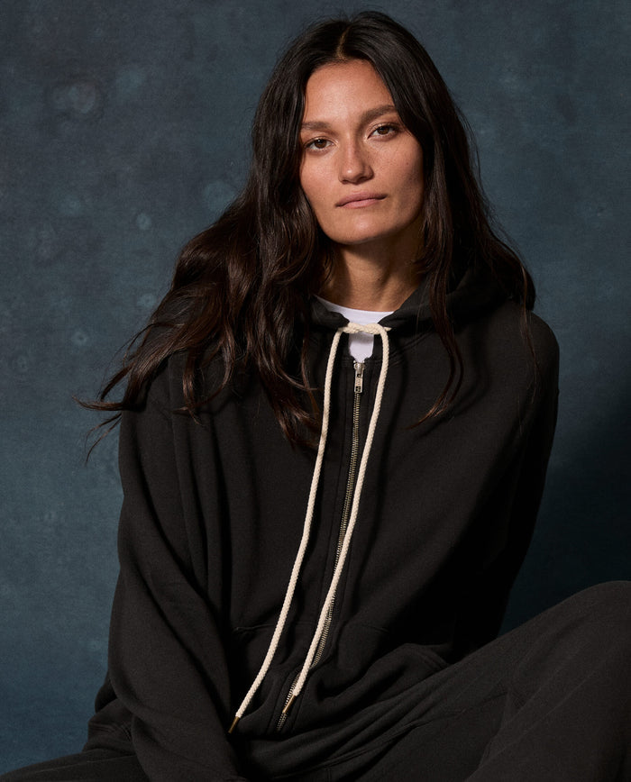 A person with long dark hair wears The Fleece Campus Hoodie in Almost Black by THE GREAT. F26 D1 FLEECE BACK KNITS, featuring white drawstrings, while sitting against a dark, textured background.