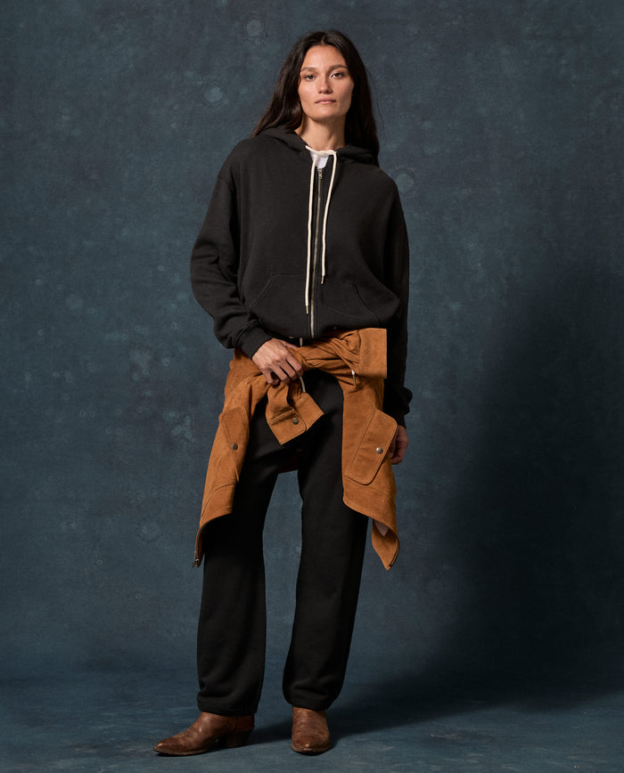 A woman stands against a dark backdrop wearing THE GREAT. F26 D1 FLEECE BACK KNITS The Fleece Campus Hoodie in Almost Black, paired with black pants, a brown shirt tied around her waist, and brown boots.