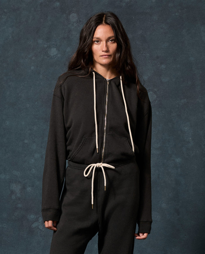 A person wearing THE GREAT. F26 D1 FLEECE BACK KNITS The Fleece Campus Hoodie in Almost Black, paired with matching black pants, stands against a dark, textured background.