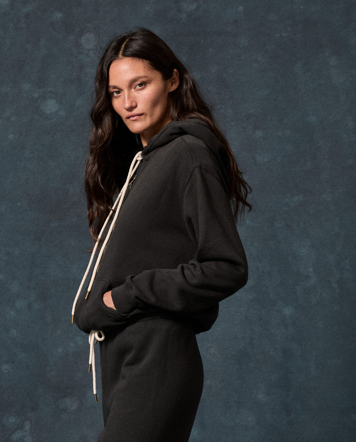 A person with long dark hair wears THE GREAT. F26 D1 FLEECE BACK KNITS The Fleece Campus Hoodie in Almost Black, featuring white drawstrings, and stands against a dark, mottled backdrop.