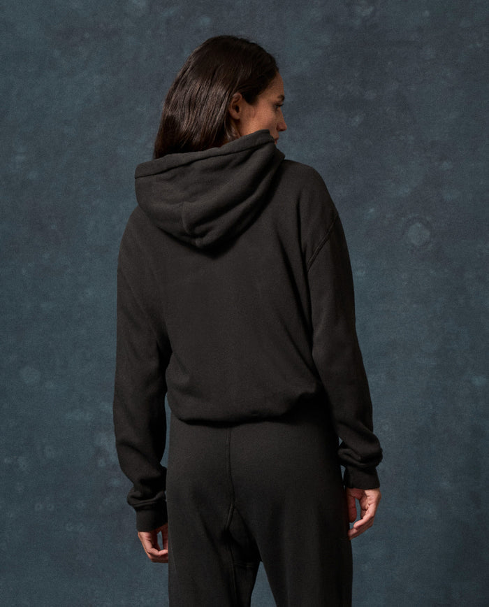 A person wearing THE GREAT. F26 D1 FLEECE BACK KNITS The Fleece Campus Hoodie in Almost Black and black pants stands with their back to the camera against a dark, textured background.