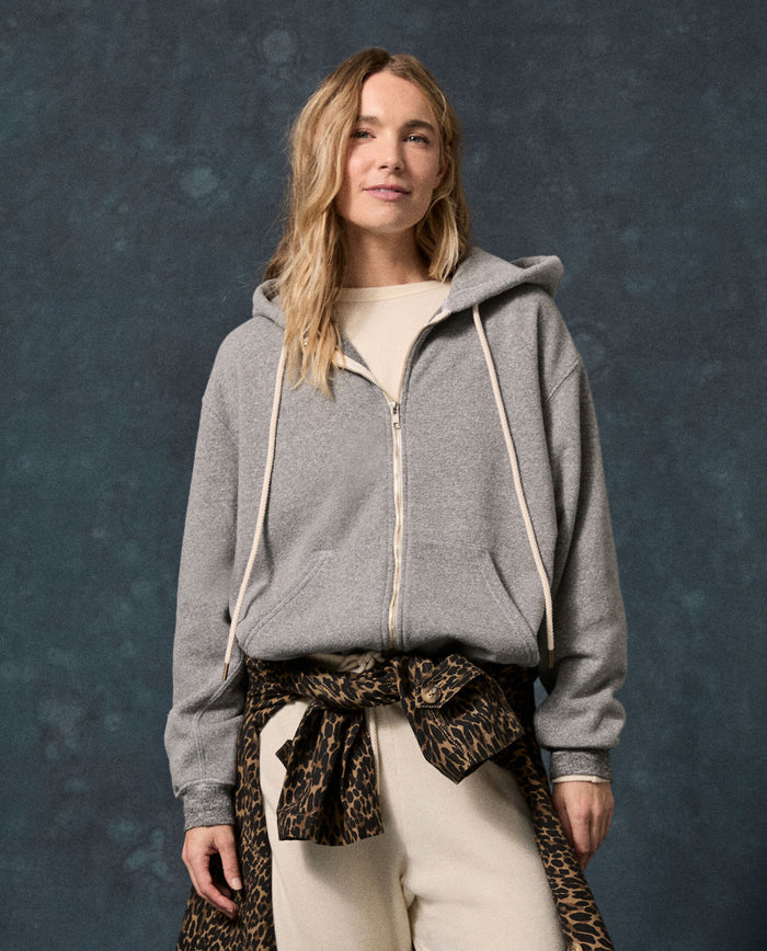 A person stands facing forward, wearing THE GREAT. F26 D1 FLEECE BACK KNITS The Fleece Campus Hoodie in Dark Heather Grey—crafted from a soft cotton-blend fleece—with a cream shirt and pants, and a leopard print jacket tied at the waist, set against a dark mottled background.
