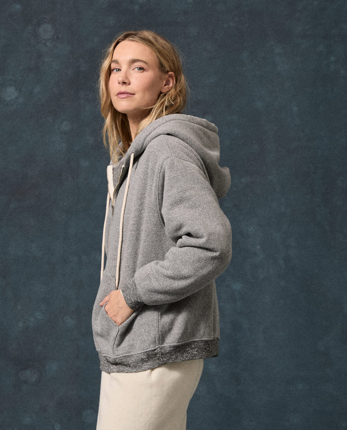 A person wears THE GREAT. F26 D1 FLEECE BACK KNITS “The Fleece Campus Hoodie” in Dark Heather Grey with light-colored pants, standing against a dark blue textured background and facing slightly to the side.