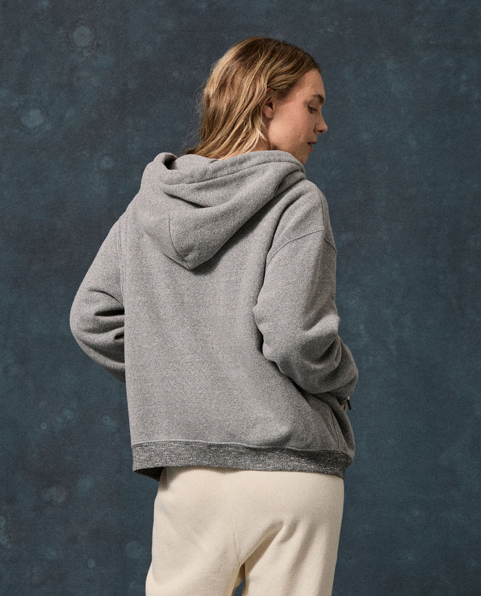 A person with long hair stands with their back to the camera against a dark textured background, wearing THE GREAT. F26 D1 FLEECE BACK KNITS’ The Fleece Campus Hoodie in Dark Heather Grey and light pants.