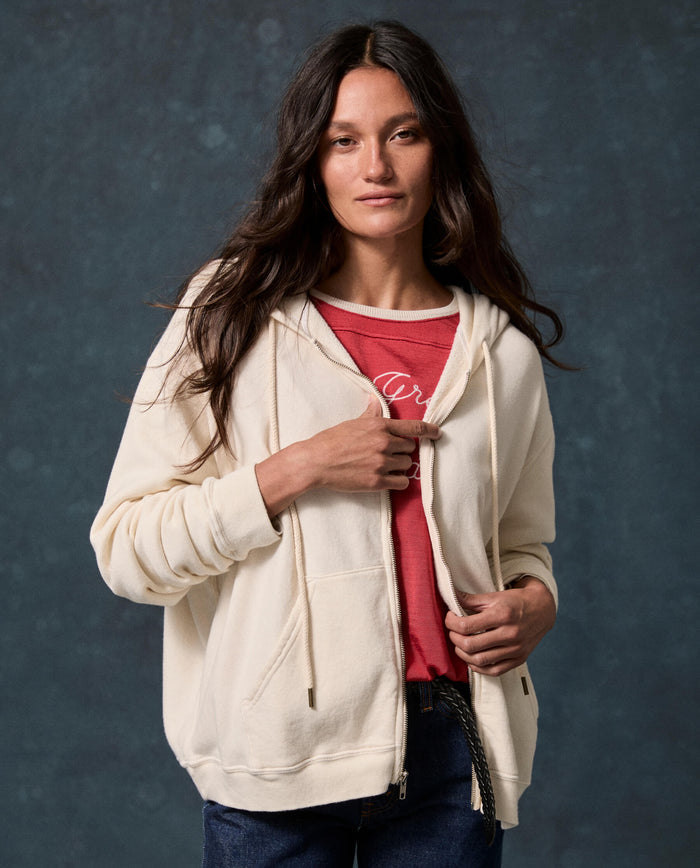 A woman with long brown hair wearing THE GREAT. F26 D1 FLEECE BACK KNITS “The Fleece Campus Hoodie” in Washed White over a red shirt and jeans, standing against a dark textured background.