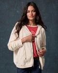 A woman with long brown hair wearing THE GREAT. F26 D1 FLEECE BACK KNITS “The Fleece Campus Hoodie” in Washed White over a red shirt and jeans, standing against a dark textured background.