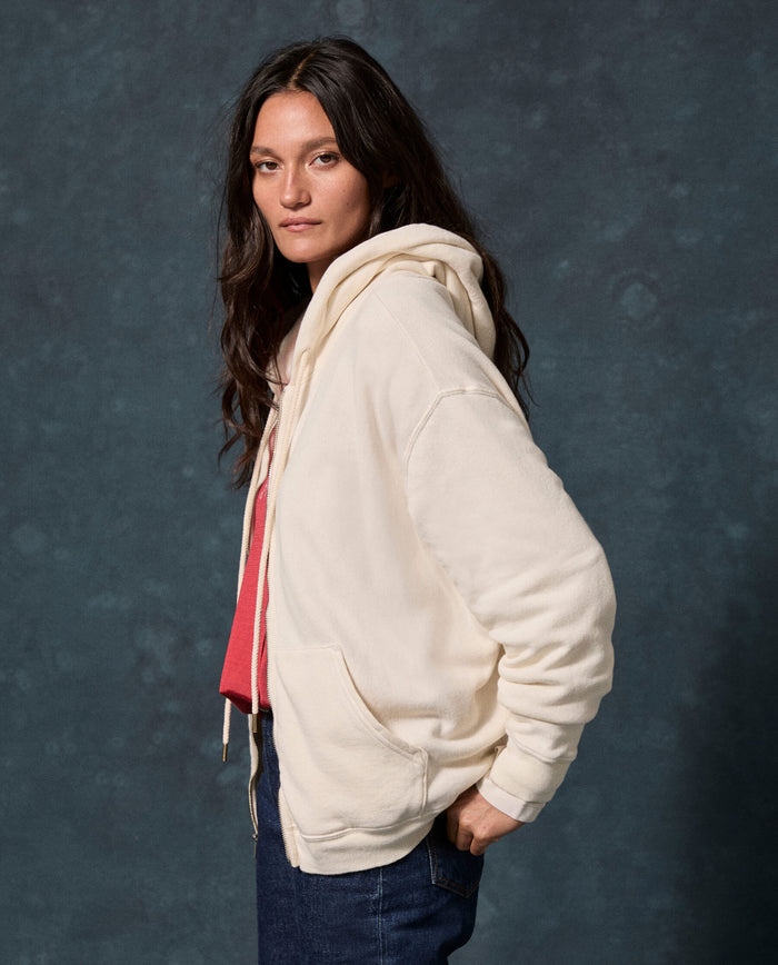 A person with long dark hair is wearing THE GREAT. F26 D1 FLEECE BACK KNITS The Fleece Campus Hoodie in Washed White over a red shirt and blue jeans, standing against a dark textured background.