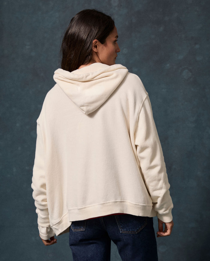 Wearing the Fleece Campus Hoodie in Washed White by THE GREAT. F26 D1 FLEECE BACK KNITS—a fleece cotton-blend zip-up sweatshirt—a person with long dark hair and dark jeans stands facing away from the camera against a dark textured background.