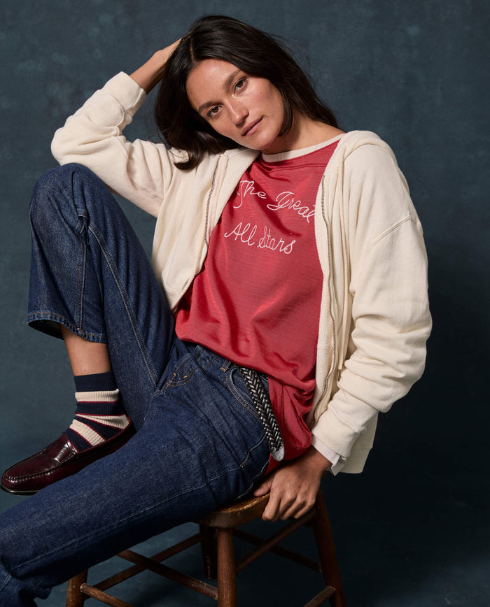 A woman sits on a stool against a dark background, wearing THE GREAT. F26 D1 FLEECE BACK KNITS The Fleece Campus Hoodie in Washed White layered over a red graphic t-shirt, with blue jeans, striped socks, and loafers. She poses with one hand behind her head.