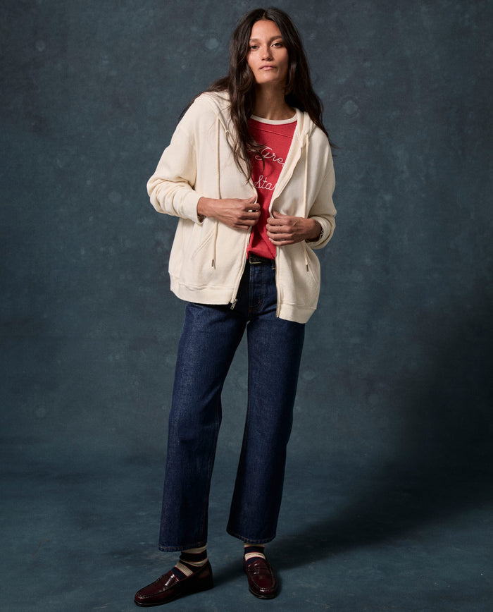 A woman stands against a dark backdrop wearing THE GREAT.’s The Fleece Campus Hoodie in Washed White—a cozy zip-up sweatshirt from the F26 D1 FLEECE BACK KNITS collection—paired with a red graphic T-shirt, blue jeans, striped socks, and brown loafers.