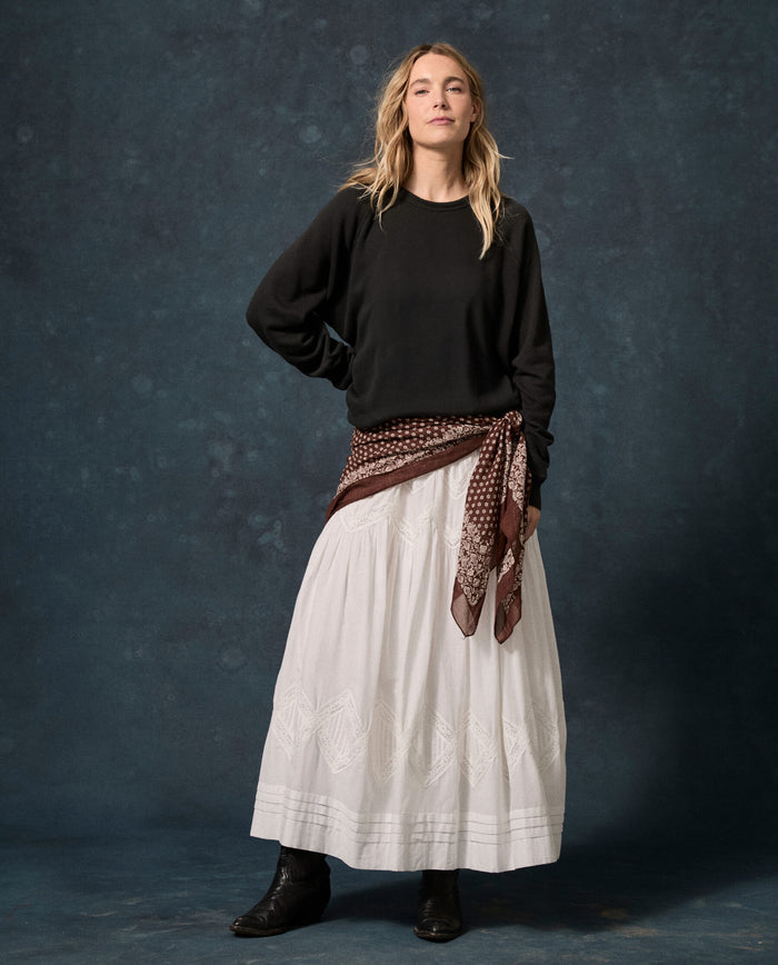 A person stands against a dark backdrop wearing THE GREAT. F26 D1 FLEECE BACK KNITS The Fleece College Sweatshirt in Solid -- Almost Black, paired with a long white skirt, black boots, and a brown patterned scarf tied at the waist.