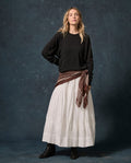 A person stands against a dark backdrop wearing THE GREAT. F26 D1 FLEECE BACK KNITS The Fleece College Sweatshirt in Solid -- Almost Black, paired with a long white skirt, black boots, and a brown patterned scarf tied at the waist.