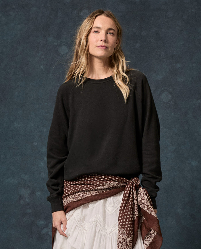 A person with long blonde hair stands in front of a textured dark blue background, wearing THE GREAT.’s The Fleece College Sweatshirt in Solid Almost Black over a white lace skirt, and accessorizing with a brown patterned scarf tied at the waist.