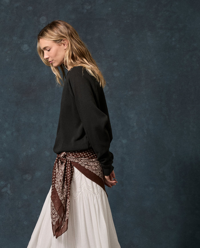 A woman in THE GREAT.’s Fleece College Sweatshirt in Solid -- Almost Black stands in profile against a dark textured background, pairing the vintage-inspired fleece-back knit top with a white skirt and a brown patterned scarf tied at her waist.