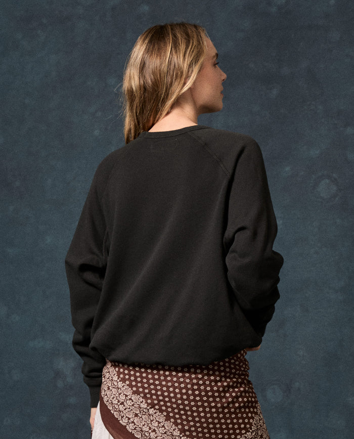 Wearing THE GREAT.’s Fleece College Sweatshirt in Solid -- Almost Black, a person with long hair and a patterned skirt stands with their back to the camera against a dark textured background.