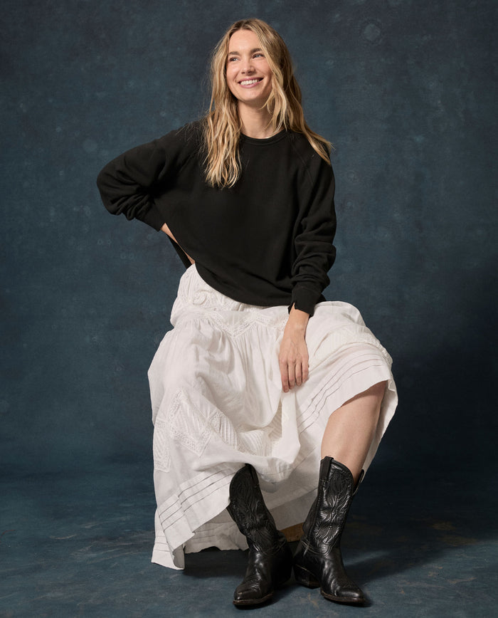 A woman sits on a stool, smiling against a dark, textured background while wearing THE GREAT. F26 D1 FLEECE BACK KNITS The Fleece College Sweatshirt in Solid -- Almost Black, paired with a long white skirt and black cowboy boots.