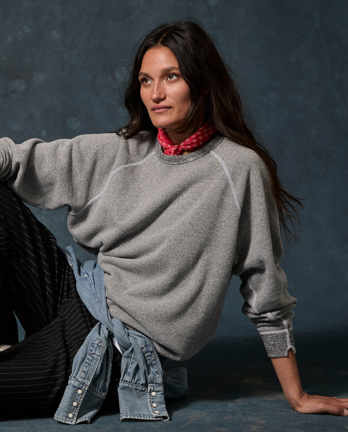 Wearing THE GREAT.’s The Fleece College Sweatshirt in Solid Dark Heather Grey, a person with long dark hair sits against a dark mottled background, styled with a red bandana, pinstriped pants, and a denim shirt tied at the waist.