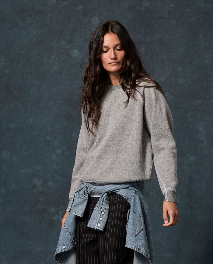 A woman with long dark hair wears THE GREAT.’s The Fleece College Sweatshirt in Solid -- Dark Heather Grey (F26 D1 Fleece Back Knits), paired with pinstriped pants and a denim shirt tied around her waist, standing against a dark mottled background.