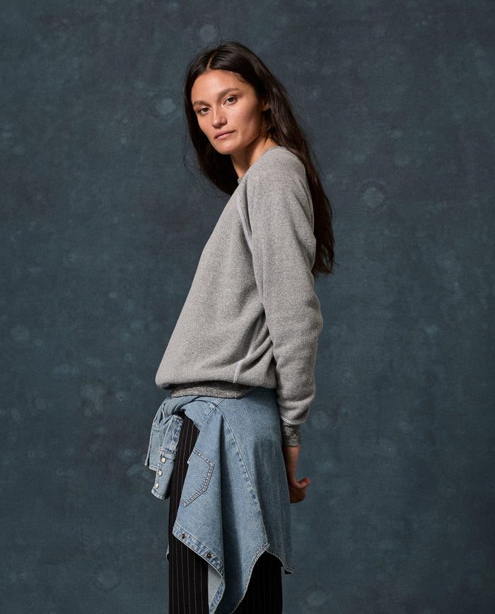 A woman with long brown hair stands sideways against a mottled dark gray background, wearing THE GREAT. F26 D1 FLEECE BACK KNITS “The Fleece College Sweatshirt” in Solid Dark Heather Grey, paired with black pants and a denim jacket tied around her waist.