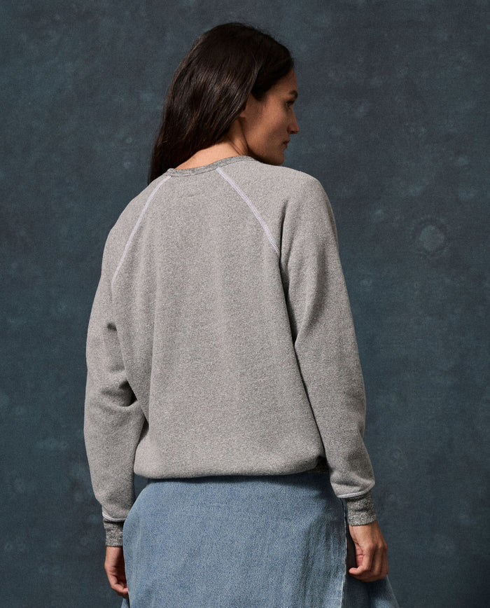 Wearing THE GREAT. F26 D1 FLEECE BACK KNITS The Fleece College Sweatshirt in Solid Dark Heather Grey, a person with long dark hair faces away from the camera in front of a dark, textured background, paired with a denim skirt.