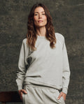 A woman with long brown hair wears THE GREAT. H25 CORE FLEECEBACK "The Fleece College Sweatshirt" in Heather Grey, standing against a textured dark background.