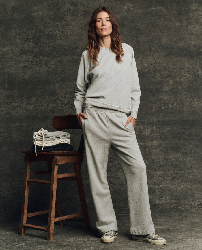 A woman poses in THE GREAT. H25 CORE FLEECEBACK The Fleece College Sweatshirt in Heather Grey, paired with sweatpants and sneakers, beside a wooden chair with folded clothes against a textured gray background.