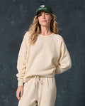 A person wearing THE GREAT. F26 D1 FLEECE BACK KNITS The Fleece College Sweatshirt in Washed White, paired with sweatpants and a green baseball cap, stands against a mottled dark background.