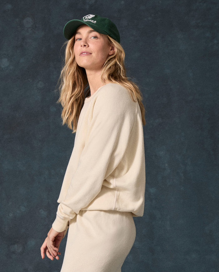Wearing THE GREAT. F26 D1 FLEECE BACK KNITS “The Fleece College Sweatshirt. Solid -- Washed White,” a person stands against a dark textured background, paired with matching pants and a green cap, gazing toward the camera.