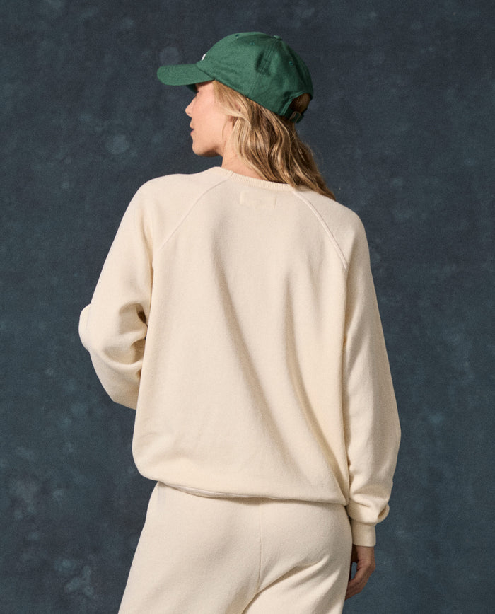 A person faces away from the camera, wearing THE GREAT.’s The Fleece College Sweatshirt in Washed White with matching pants and a green baseball cap, set against a dark mottled background.