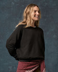 A person with long, wavy hair wears THE GREAT. The Fleece Rookie Sweatshirt in Solid -- Almost Black (F26 D1 FLEECE BACK KNITS), paired with a red patterned garment tied around the waist, standing against a dark mottled background.