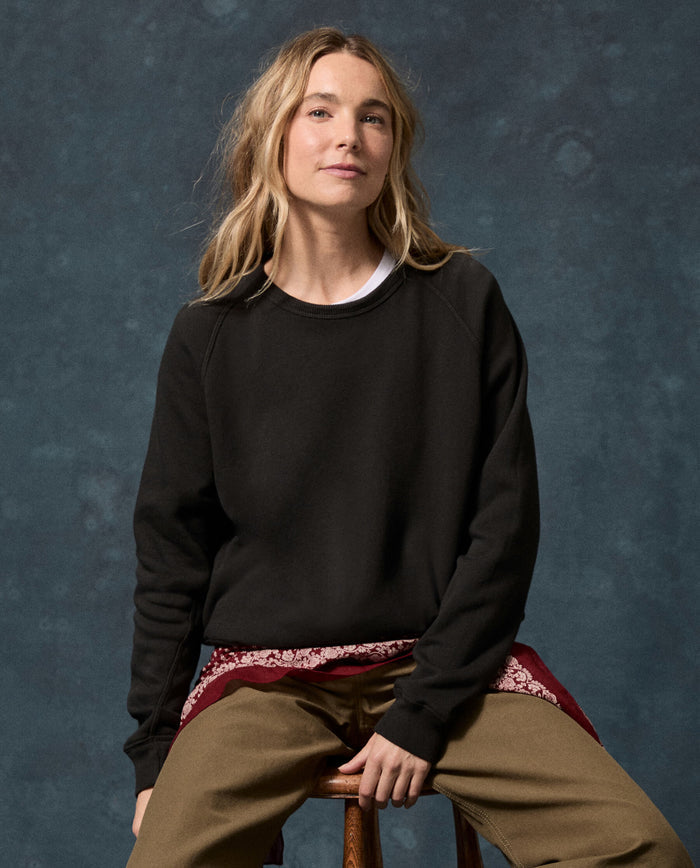 A person with long blond hair wearing THE GREAT. The Fleece Rookie Sweatshirt in Solid -- Almost Black from the F26 D1 FLEECE BACK KNITS collection, paired with a white shirt, brown pants, and a red patterned scarf, sits on a stool against a dark blue textured background. This cozy sweatshirt is made in Los Angeles.